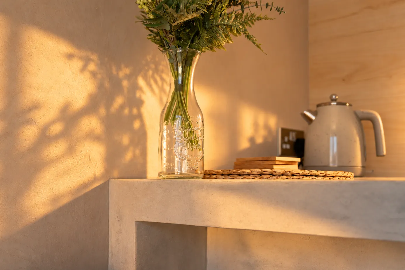 Shadow of flowers on a plaster wall beside a glass vase and kettle in golden hour light